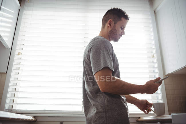Чоловік використовує свій мобільний телефон, тримаючи чашку кави на кухні вдома — стокове фото