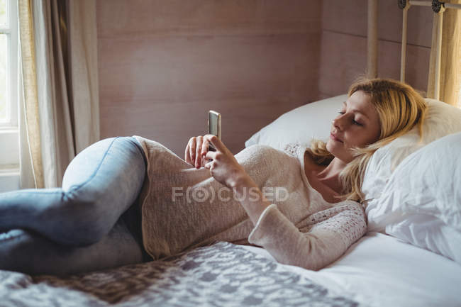 Женщина лежит на кровати и пользуется мобильным телефоном в спальне дома — стоковое фото