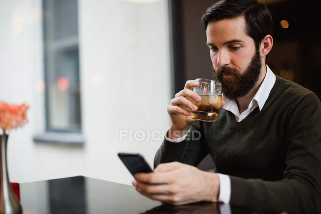 Hombre tomando un vaso de bebida mientras usa el teléfono móvil en el bar - foto de stock