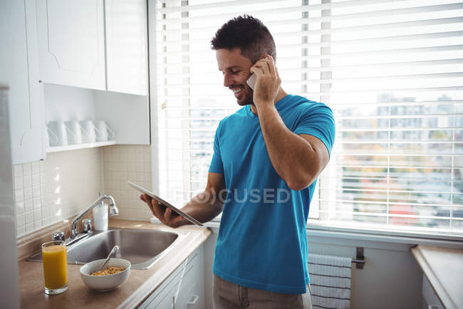Чоловік розмовляє на мобільному телефоні, використовуючи цифровий планшет на кухні вдома — стокове фото