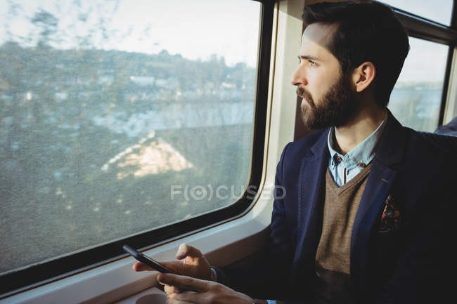 Young businessman holding mobile phone and looking through train window — Stock Photo
