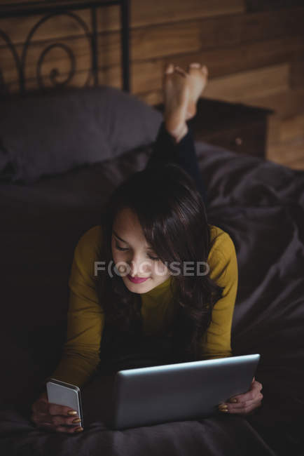 Donna sdraiata sul letto con tablet digitale e telefono cellulare in camera da letto a casa — Foto stock