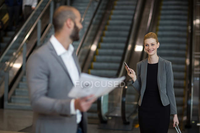 Улыбающаяся деловая женщина, взаимодействующая с бизнесменом в зоне ожидания в терминале аэропорта — стоковое фото