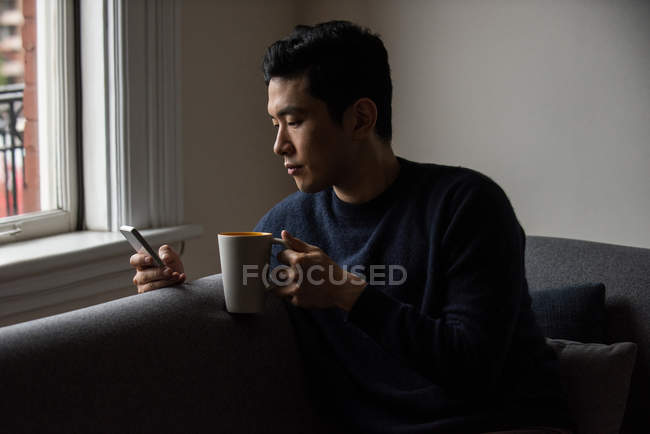Homme utilisant un téléphone portable tout en prenant une tasse de café à la maison — Photo de stock