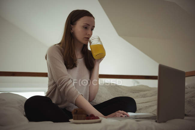 Donna che ha succo mentre usa il computer portatile sul letto a casa — Foto stock
