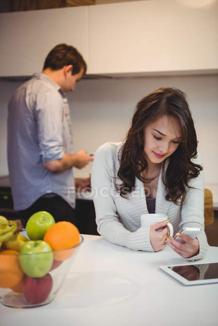 Mulher usando telefone celular, enquanto o homem trabalhando no fundo na cozinha — Fotografia de Stock