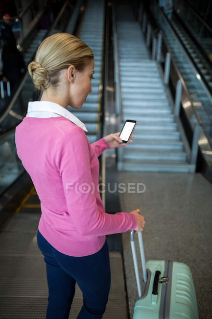 Пригородная женщина с багажом с помощью мобильного телефона на эскалаторе в аэропорту — стоковое фото