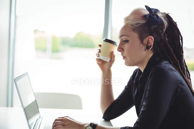Женщина держит одноразовый кофе чашку во время использования ноутбука в кафе — стоковое фото