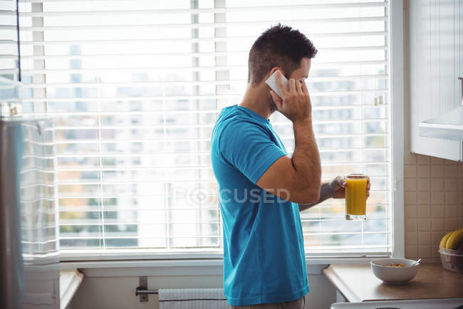 Homem falando no telefone celular ao usar ter vidro de suco na cozinha em casa — Fotografia de Stock