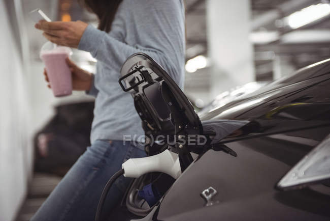 Car being charged with electric car charger while woman standing in background at electric vehicle charging station — Stock Photo