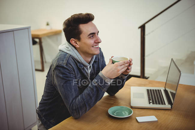 Улыбающийся мужчина пьет кофе с ноутбуком на столе в кафе — стоковое фото