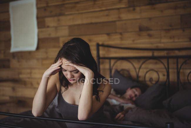 Расстроенная женщина сидит на кровати, в то время как мужчина спит на заднем плане в спальне — стоковое фото
