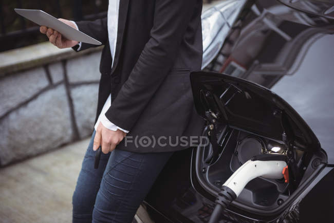 Partie médiane de la femme utilisant une tablette numérique lors de la charge de la voiture électrique à la station de charge du véhicule — Photo de stock