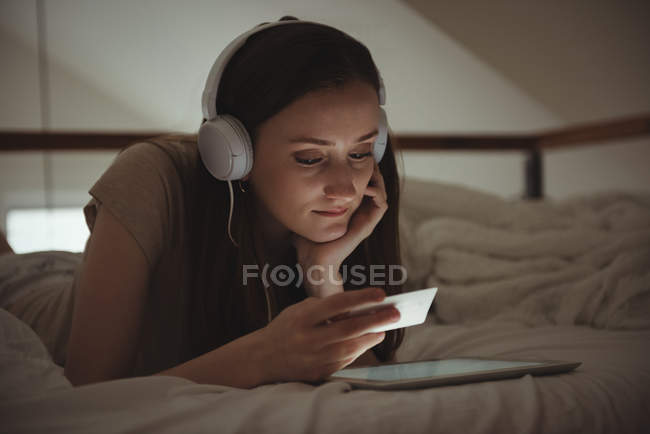 Mulher com fones de ouvido olhando para o cartão ao usar tablet digital na cama — Fotografia de Stock