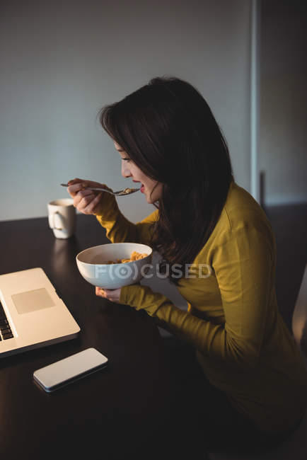 Mujer comiendo cereales mientras trabaja en el ordenador portátil en la sala de estudio en casa - foto de stock