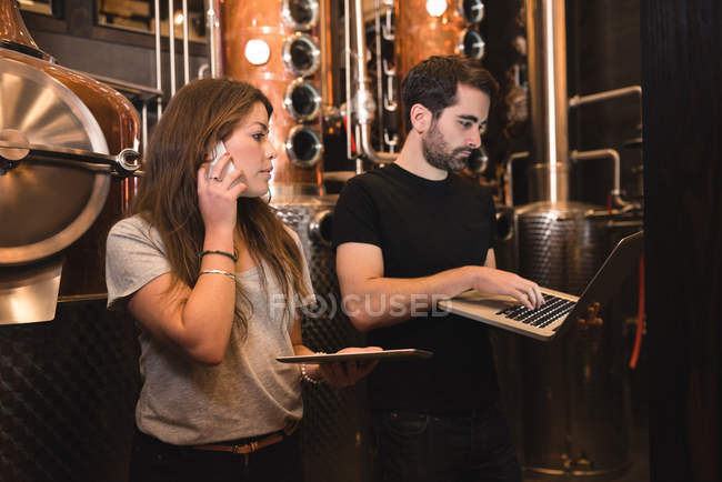 Homme utilisant un ordinateur portable tandis que la femme parle au téléphone dans l'usine de bière — Photo de stock