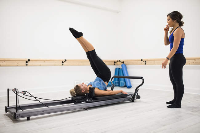 Entrenadora mujer que ayuda a la mujer con el ejercicio de estiramiento en reformador en el gimnasio - foto de stock