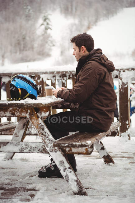 Mann auf seinem Handy sitzt an einem Tisch draußen in einem Skigebiet — Stockfoto