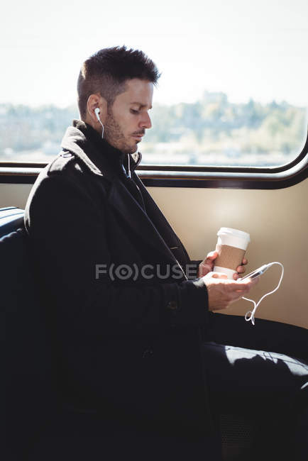 Empresario sosteniendo una taza de café desechable y escuchando música en el teléfono móvil en el tren - foto de stock