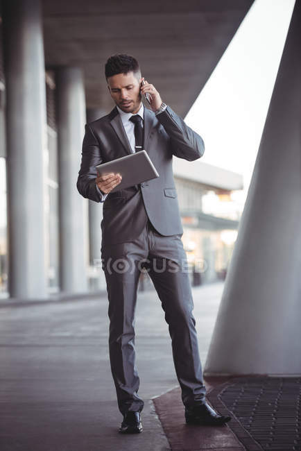 Businessman using digital tablet while talking on mobile phone in the office campus — Stock Photo