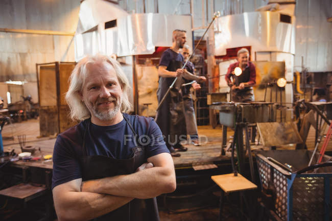 Портрет стеклодува, стоящего со скрещенными руками, в то время как коллеги работают на заднем плане на стекольном заводе — стоковое фото
