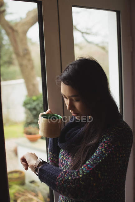 Женщина смотрит на умные часы, когда пьет кофе дома — стоковое фото