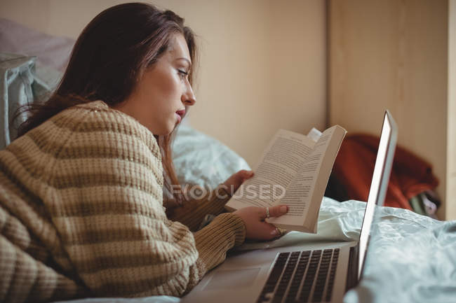 Beautiful woman reading a book and using laptop on bed at home — Stock Photo