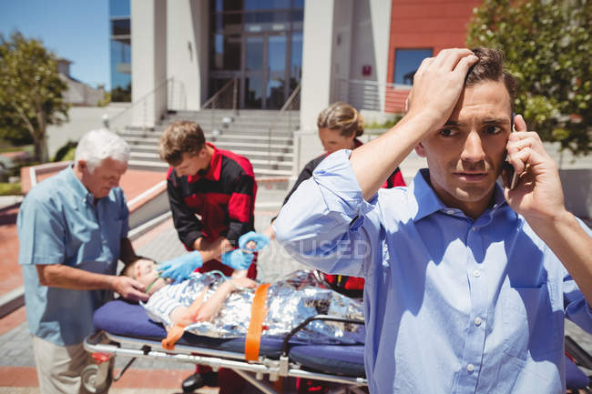 Man talking on mobile phone and paramedics examining injured boy on street in background — Stock Photo