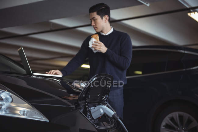 Homme utilisant un ordinateur portable tout en chargeant une voiture électrique dans le garage — Photo de stock