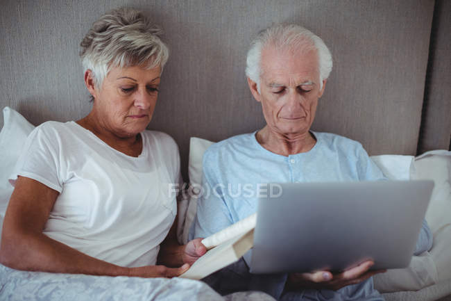 Mujer mayor leyendo libro mientras que el hombre mayor usando el ordenador portátil en la cama en la habitación - foto de stock