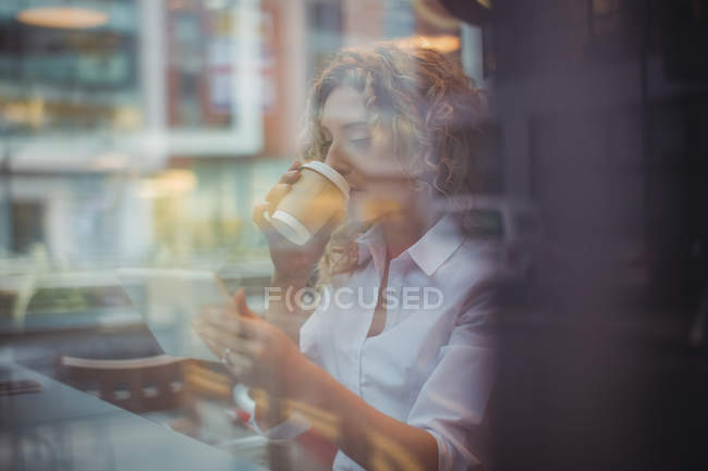 Mid adult businesswoman drinking coffee and using phone at counter in cafeteria — Stock Photo