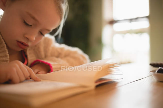 Mädchen im Grundschulalter sitzt am Tisch und liest zu Hause Buch — Stockfoto