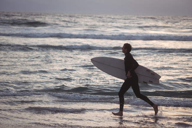 Вид сбоку на человека с доской для серфинга, бегущего по пляжу в сумерках — стоковое фото