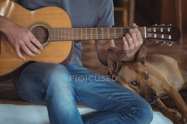 Sección media de un hombre tocando la guitarra en casa, perro acostado a su lado - foto de stock