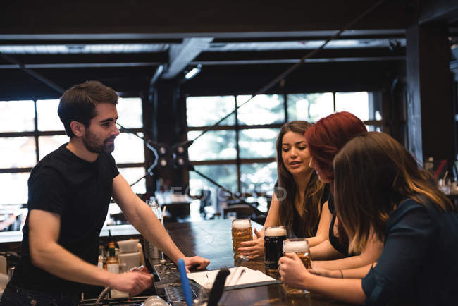 Amigos segurando copos de cerveja no balcão do bar e interagindo com o barman — Fotografia de Stock