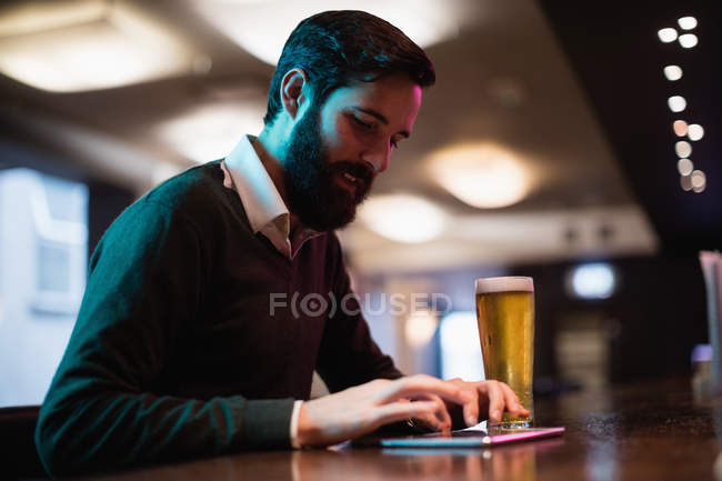 Человек, использующий цифровой планшет со стаканом пива на стойке в баре — стоковое фото