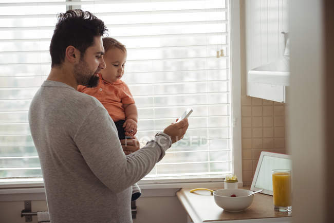 Чоловік середнього віку, який використовує мобільний телефон, тримаючи сина на кухні вдома — стокове фото