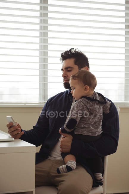 Батько тримає дитину, використовуючи мобільний телефон за столом вдома — стокове фото