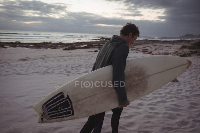 Homem carregando prancha de surf andando na praia ao entardecer — Fotografia de Stock