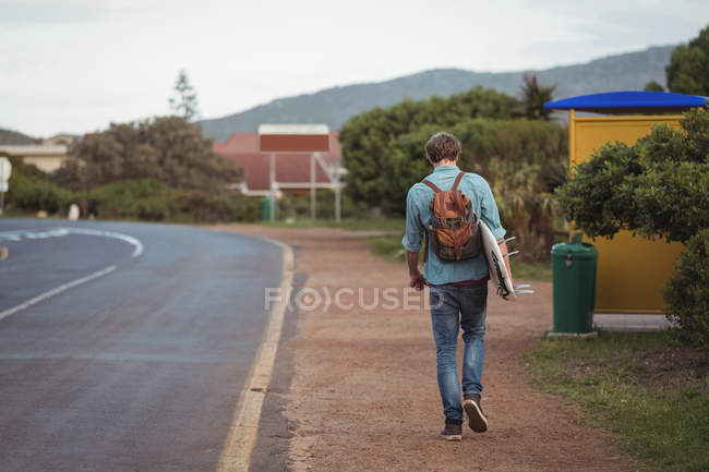 Visão traseira do homem com mochila carregando uma prancha de surf andando ao longo da estrada — Fotografia de Stock