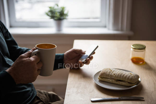 No meio da seção do homem usando telefone celular enquanto toma uma xícara de café em casa — Fotografia de Stock