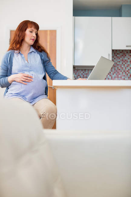 Беременная женщина использует ноутбук на кухне дома — стоковое фото