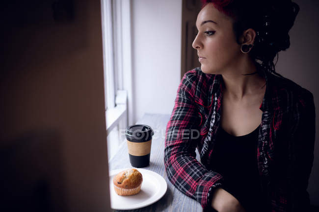 Продумана жінка дивиться крізь вікно з кексом і чашкою кави на столі в кафе — стокове фото