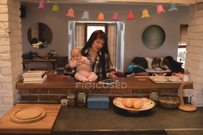 Mother using digital tablet while carrying baby in kitchen at home — Stock Photo