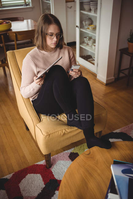 Mulher sentada na cadeira usando telefone celular e tablet digital na sala de estar — Fotografia de Stock