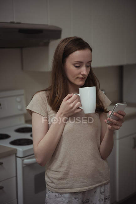 Frau trinkt Kaffee, während sie ihr Handy zu Hause in der Küche benutzt — Stockfoto