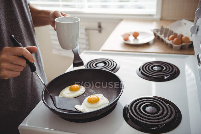Homme avec tasse à café utilisant spatule pour la cuisson des œufs frits dans la cuisine — Photo de stock