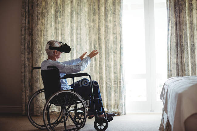 Homem sênior sentado em cadeira de rodas e usando fone de ouvido realidade virtual no quarto em casa — Fotografia de Stock