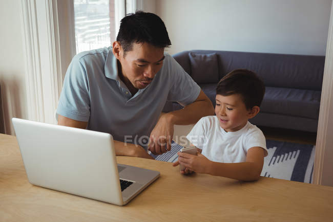 Padre e hijo usando teléfono móvil y portátil en la sala de estar en casa - foto de stock