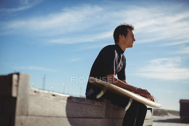 Nachdenklicher Surfer sitzt auf einer Holzplattform mit Surfbrett am Strand — Stockfoto